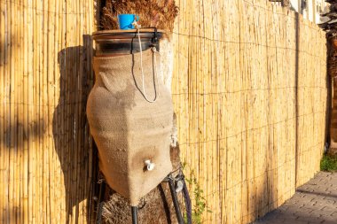 Fas, Tangier 'de bir sokakta, kuru kamışların arka planında palmiye ağacına bağlanmış plastik mavi bardakta su içilen kil testisi.