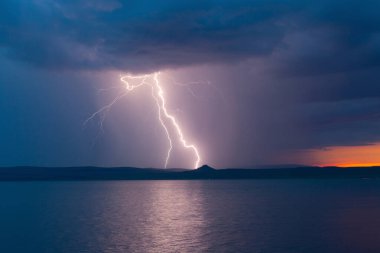 Alacakaranlık fırtınalı gökyüzünde, göl suyunun üzerinde bir gök gürültüsü sırasında parlak bir şimşek çakar.