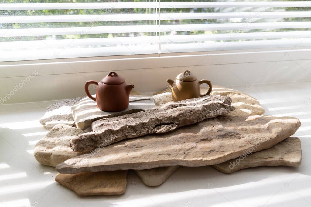 Two brown asian teapots for a tea ceremony stand on stone slabs on a white windowsill