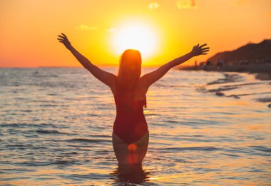 Okyanusun arka planında mayo giymiş bir kadının siyah silueti. Deniz altyapısında mutlu genç bir kadın gün batımında elleri açık özgürlüğün tadını çıkarıyor.