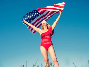 Kumsalda Amerikan bayrağı tutan güzel, neşeli sarışın kadın. Kırmızı bikinili orta yaşlı bir kadın vatansever bir şekilde coşkuludur. Seyahat et. ABD bayrağı