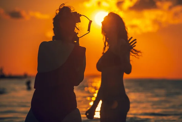 Black silhouettes of mom and daughter during a professional photo shoot at sunset. A beautiful brunette model in a swimsuit being a photographer. The focus is on the photographer