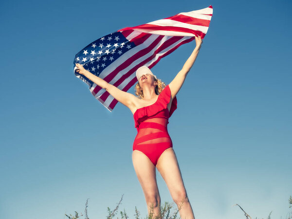 Красивая веселая блондинка с американским флагом на пляже. Женщина средних лет в красном бикини патриотически увлечена. Путешествие. Флаг США