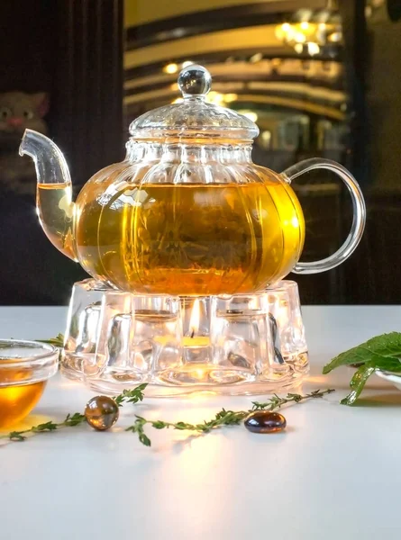 白いテーブルの上のキャンドルに温まるガラスのティーポットで香り高い新鮮なハーブティーとメリッサ 伝統的なハーブドリンク 蜂蜜とお茶 ケトル暖かい — ストック写真