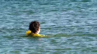 Koyu renk saçlı küçük bir çocuk şişme yelekle denizde yüzüyor.