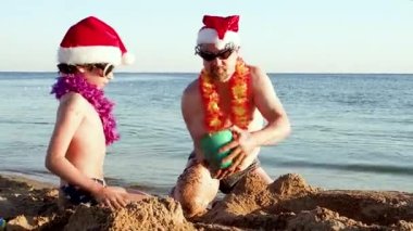 Baba ve küçük oğlu Noel Baba 'nın şapkasıyla kumdan kale yapıp kumsalda oynuyorlar.