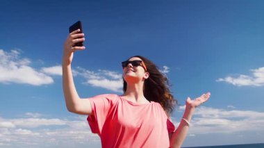 Mavi gökyüzü arka planında dururken cep telefonu kullanan genç ve güzel bir kadın.