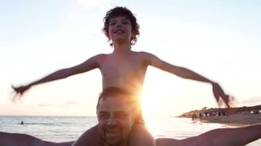 Oğlu babasının omuzlarında oturur ve gün batımında uçuşu taklit ederek el kaldırır. Deniz kenarında bir aile. Boşluğu kopyala