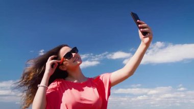 Mavi gökyüzü arka planında dururken cep telefonu kullanan genç ve güzel bir kadın.