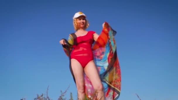 美しい陽気なブロンドの女性がビーチでスカーフを持っています ビーチで中年の女性 赤いスカーフを持った女性が風に乗って飛んでいる 水着の女の子は砂の島で夏休みをお楽しみください — ストック動画