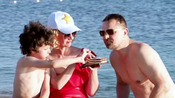 日没の間にビーチに座っている3人の家族スポンジケーキを食べる — ストック動画