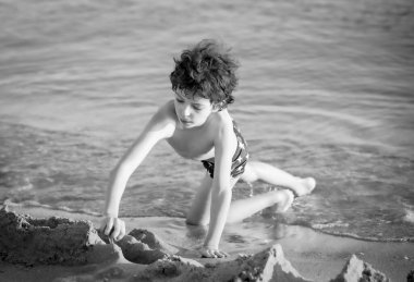 Tatlı kıvırcık erkek çocuk sahilde oyun oynuyor. Küçük çocuk yaz tatilinde kumla oynuyor. Yaz tatilinde komik oyunlar oynuyor. Çocuk sahilde eğleniyor. Boş zaman, oyunlar ve tatil. Siyah beyaz fotoğraf.