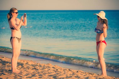Anne kızının tropik plajda akıllı telefon kullanarak fotoğrafını çekiyor.