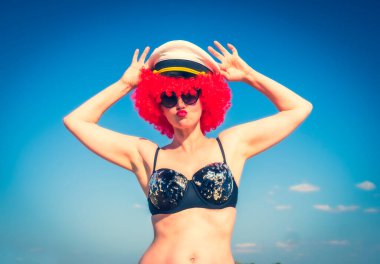 Kırmızı peruklu, orta yaşlı deli bir kadının portresi. Siyah, parlak bir mayo. Dudaklarını gökyüzüne karşı bir öpücük için uzattı. Tropik bir tatil köyünde orta yaşlı bir kadın.