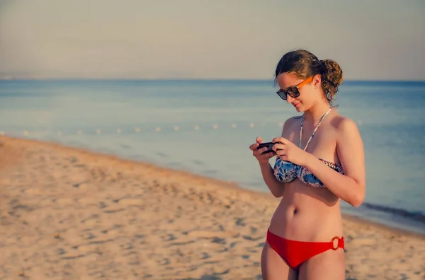 漂亮的少女黑发使用智能手机站在海滩上 背景是大海和地平线 复制空间 — 图库照片