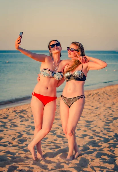 母亲和女儿在海滩上笑着做鬼脸 在海滩上 妈妈和年轻的黑发姑娘们正在用手机拍照 — 图库照片