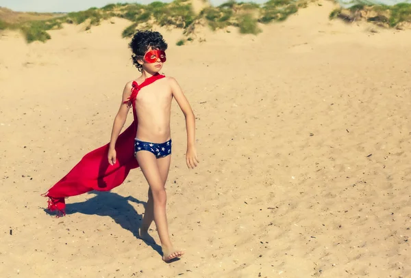 Little 9 year old child superhero with red cloak and glasses. Boy superhero on beach in summer vacation. Copy space