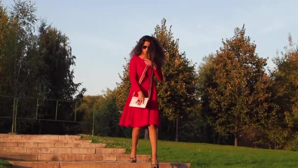 ヒールのエレガントな女性と赤いドレスは卒業後に街の公園を歩いていて 横から横にずれています — ストック動画