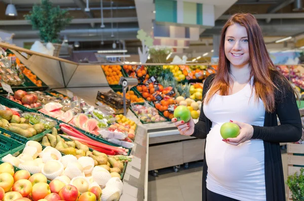 Беременная женщина с зеленым яблоком выбирает овощи в супермаркете — стоковое фото