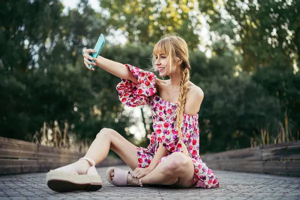 夏天坐在人行道上 拿着智能手机自拍的笑容满面的年轻女子的画像 — 图库照片