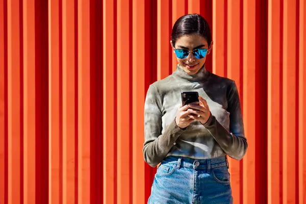 晴れた日に赤い段ボールの壁に立っている間 スマートフォンを使用して若い女性を笑顔 — ストック写真