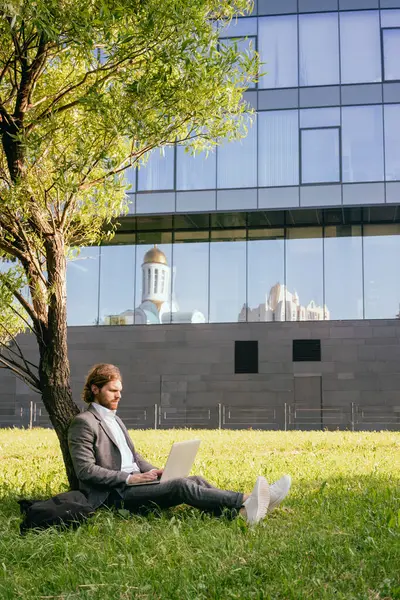 市中心 年轻的男性创业者坐在草地上 一边用笔记本电脑 一边坐在树上 — 图库照片