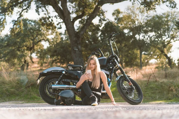 Блондинка Байкер Помощью Смартфона Сидя Против Мотоцикла Обочине Дороги — стоковое фото