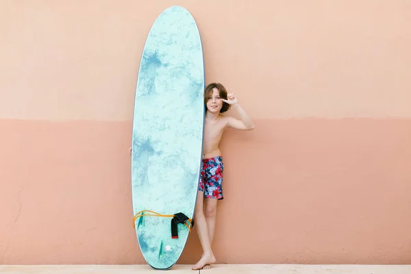 Junge Badehose Gestikuliert Während Mit Surfbrett Vor Beiger Wand Steht — Stockfoto