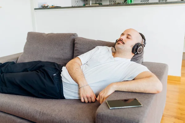 家でソファでリラックスしながら ヘッドフォンを介して音楽を聞き閉じ目を持つ大胆な男 — ストック写真