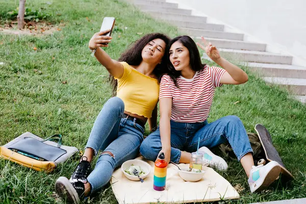 年轻女子坐在公园里 一边和女友用手机自拍 一边示意和气 — 图库照片