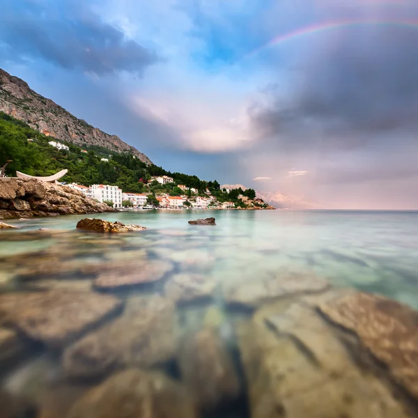 Arcobaleno sulla spiaggia rocciosa e piccolo villaggio dopo la pioggia — Foto Stock