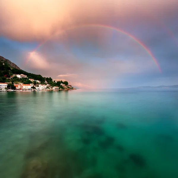 Arcobaleno sulla spiaggia rocciosa e piccolo villaggio dopo la pioggia — Foto Stock