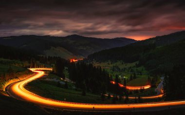 Araba ışığı, dağların kıvrımlı yolunda çarpıcı bir gökyüzü ile yol alır.