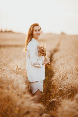 Uzun kıvırcık saçlı ve açık beyaz takım elbiseli genç bir kız buğday tarlasında kameraya poz veriyor, günbatımının arka planında.