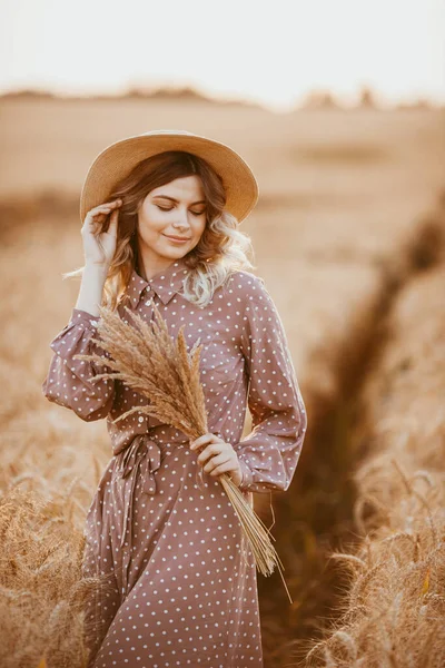 白い水玉の茶色のドレスを着た若い女の子が 夕日を背景に小麦畑に立っています — ストック写真