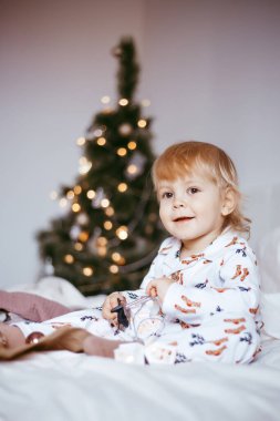 Çocuğun ilk Noel 'i. Düşünceli küçük bir çocuk, altın renkli çelenkli, parlak bir köknar ağacının arka planında, tatil pijamalarıyla yatıyor. Noel konsepti.