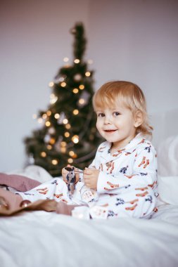 Çocuğun ilk Noel 'i. Düşünceli küçük bir çocuk, altın renkli çelenkli, parlak bir köknar ağacının arka planında, tatil pijamalarıyla yatıyor. Noel konsepti.