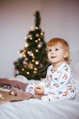 Çocuğun ilk Noel 'i. Düşünceli küçük bir çocuk, altın renkli çelenkli, parlak bir köknar ağacının arka planında, tatil pijamalarıyla yatıyor. Noel konsepti.