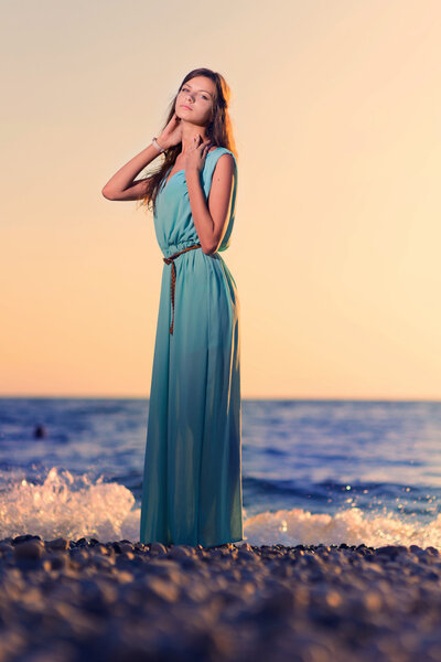 Young woman in dress on beach at sunset against sun