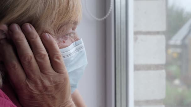Anciana en máscara protectora mira por la ventana y sostiene su cabeza enferma en las manos — Vídeos de Stock