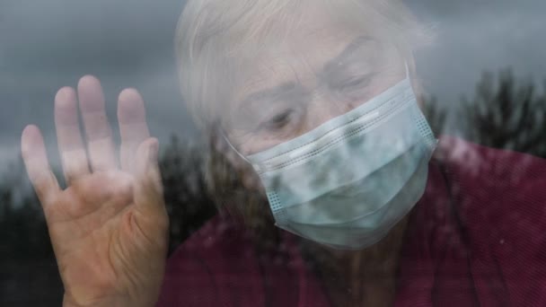 Eine ältere Frau mit Maske im Gesicht blickt aus dem Fenster auf der Straße — Stockvideo