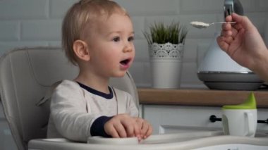 Anne, bebek koltuğunda oturan çocuğuna kaşıkla yulaf lapası veriyor.