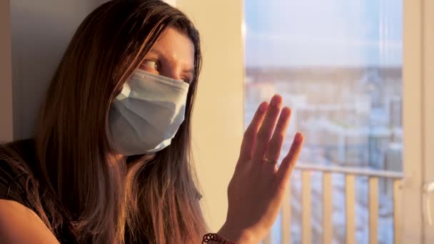 Joven mujer caucásica en máscara se ve con suerte por la ventana al atardecer — Vídeos de Stock