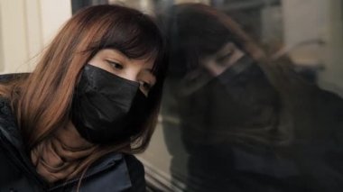 Virüs maskesi takan üzgün kadın metroya biner ve pencereden dışarı bakar.