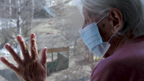 Vieille femme en masque médical sur le visage regarde la rue avec sa main sur le verre — Video