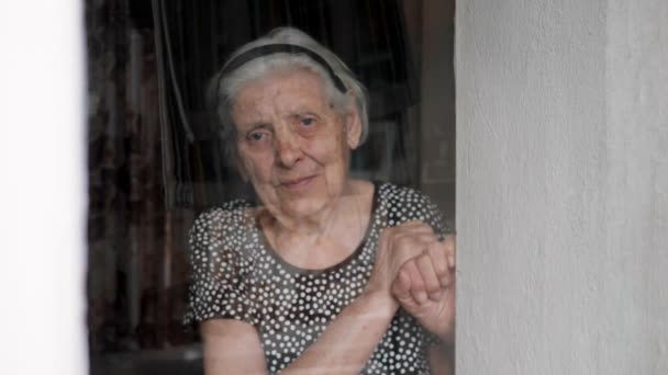 Triste anciana mujer se para en la ventana y mira en la cámara en cuarentena — Vídeos de Stock