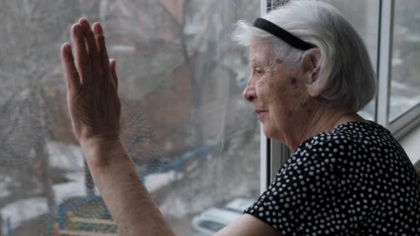 Stara babcia wygląda spokojnie przez okno z ręką na szkle. — Wideo stockowe