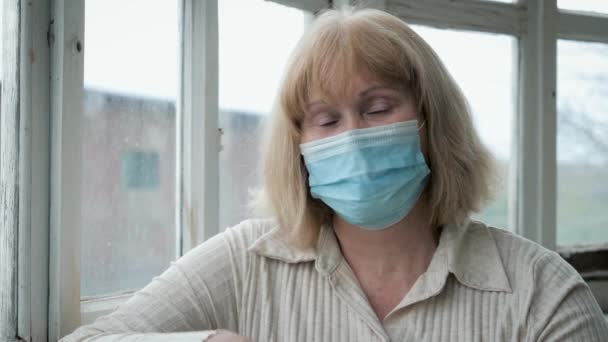 Portræt af moden kvinde i medicinsk maske på hendes ansigt ved vinduet – Stock-video