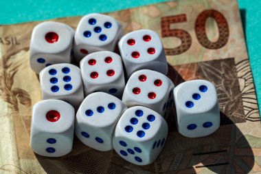 50 Brezilya banknotuna dağılmış kırmızı ve mavi noktalı birkaç beyaz zarın yakın çekimi. Kompozisyon kumar ve para arasındaki zıtlığı vurguluyor ve bahis, mali risk ve şans gibi konuları ortaya koyuyor. Işıklandırma vurgulanıyor
