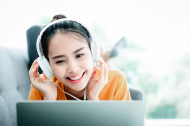 Laptop kullanan, kulaklık takan, müzik dinleyen ve oturma odasında kanepede uzanan güzel Asyalı bir kadın.